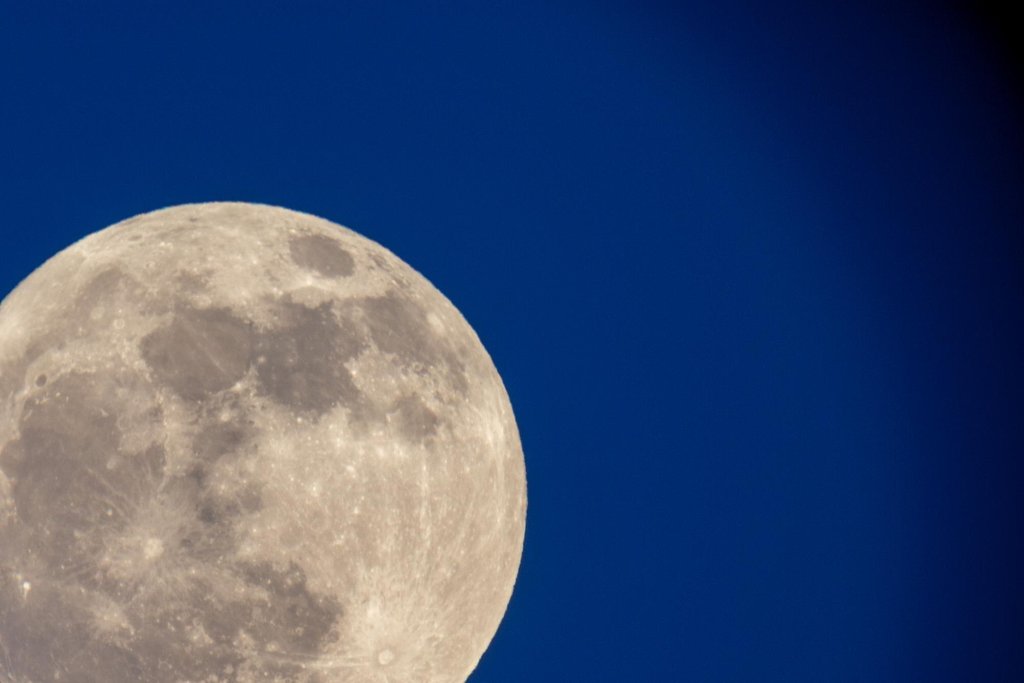 A nearly full Moon photographed against a deep blue daytime sky, craters and dark maria visible on the lit disk.