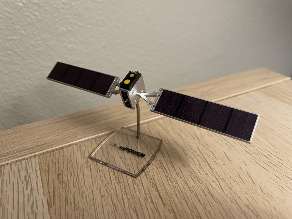A tabletop scale model of an O3b mPOWER satellite with two solar arrays extended, standing on a clear base engraved O3b mPOWER.