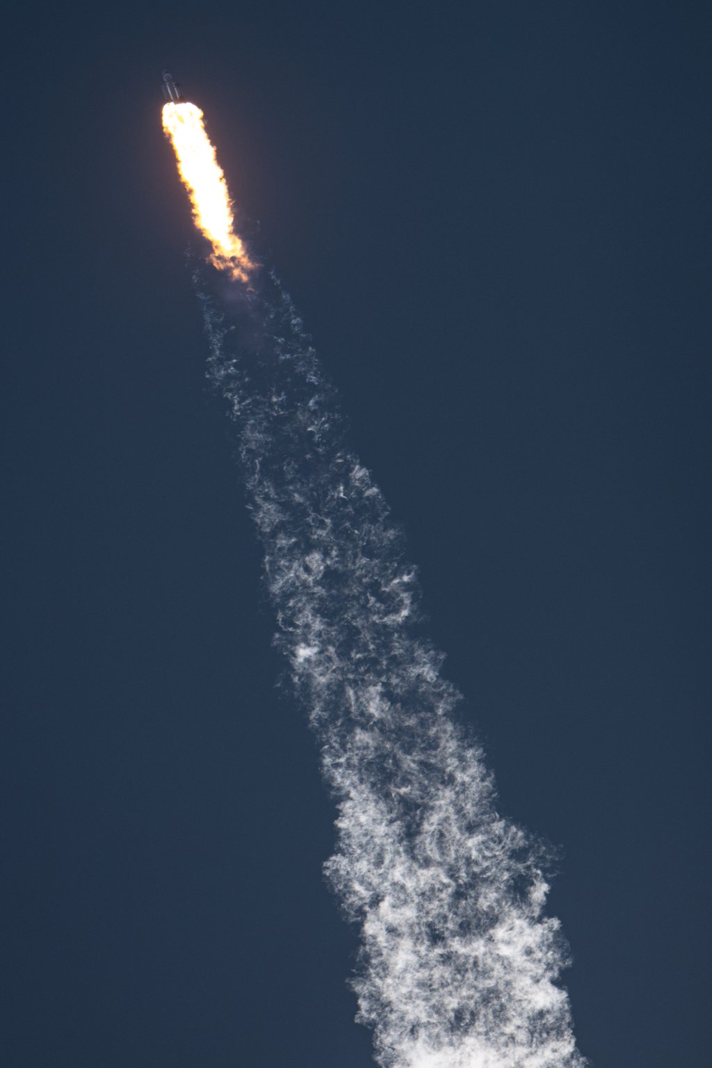 A rocket arcing across a deep blue sky, trailing a long curled exhaust plume.