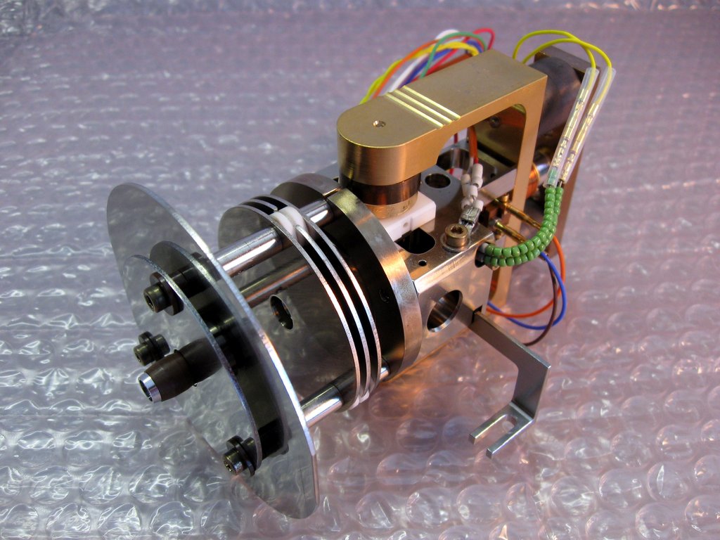 The metal ion source assembly of a mass spectrometer, a cylinder of machined steel with colored wiring, photographed on a bench.