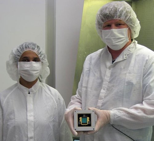 Two engineers in clean-room suits, hairnets and masks hold up a small square detector module for the MIRI instrument.