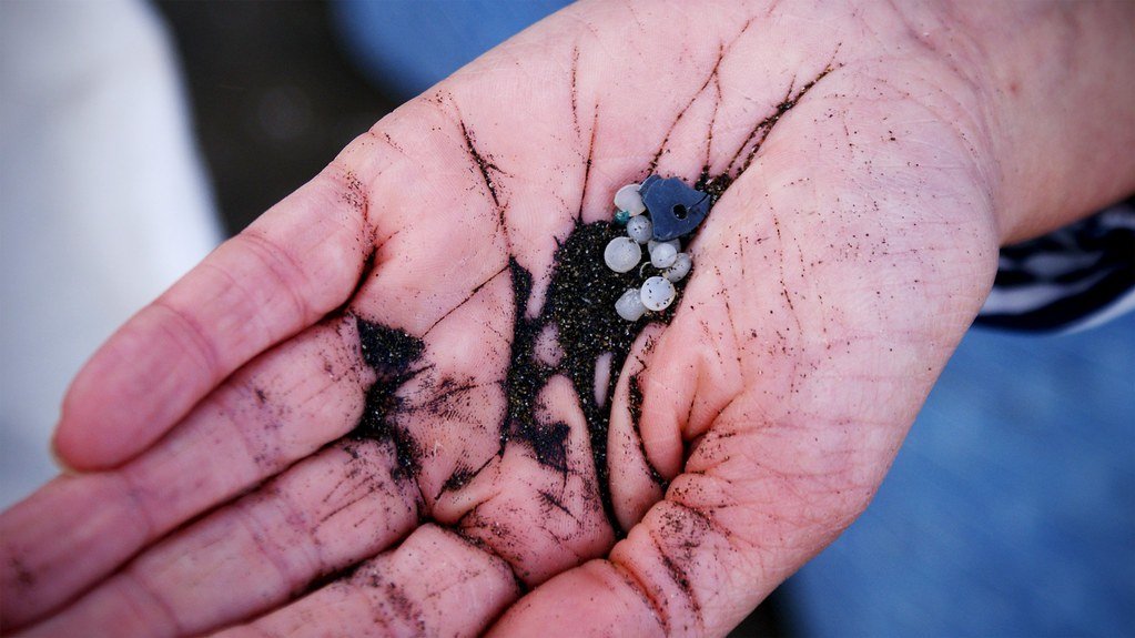 A cupped hand holding white plastic pellets and one blue plastic fragment mixed with dark sand.