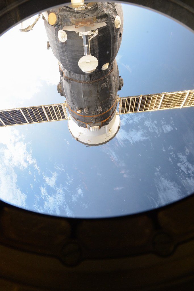 A Progress cargo ship docked to the International Space Station, seen through a round station window with Earth below.