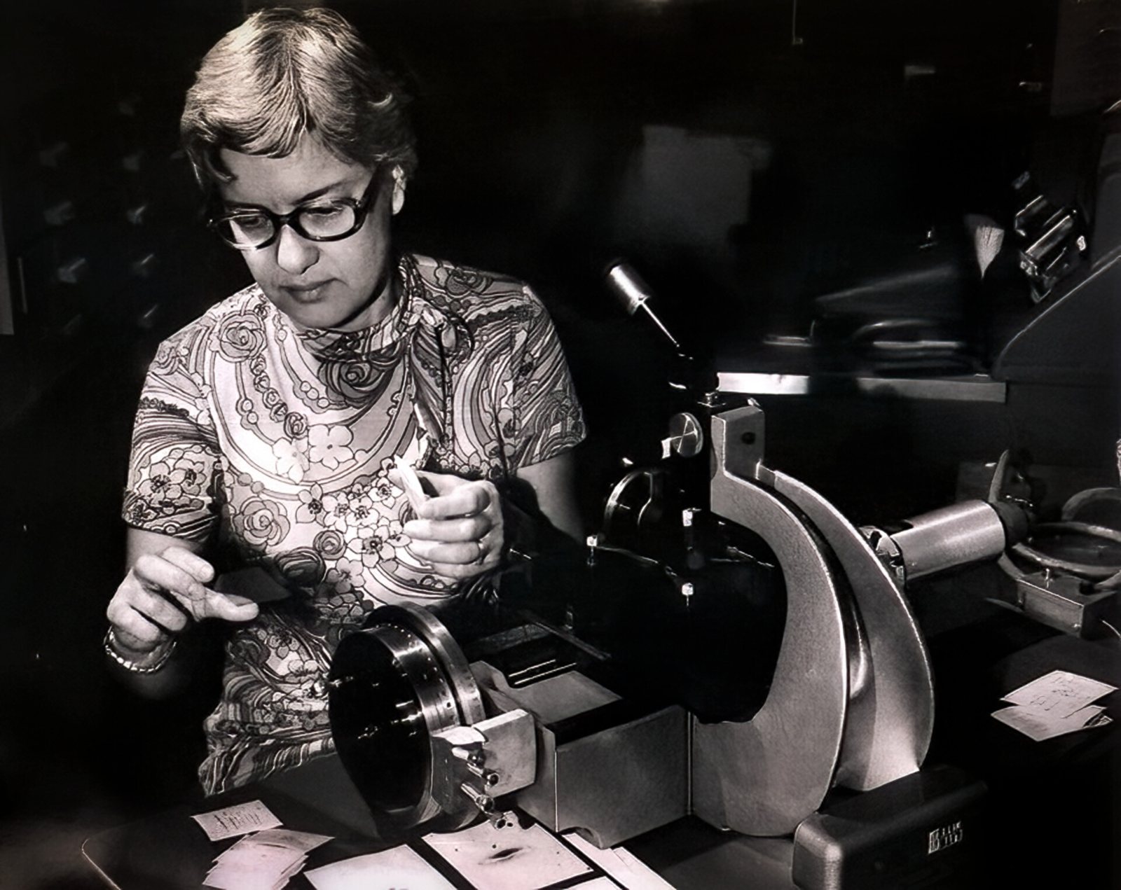 Black-and-white photograph of Vera Rubin in 1974, seated at a plate-measuring machine, holding a photographic plate, with galaxy spectra laid out on the table.