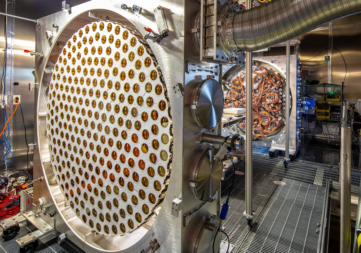 A large circular array of several hundred gold-coloured photomultiplier tubes mounted in a white holder, standing in a laboratory beside stainless-steel vacuum hardware.