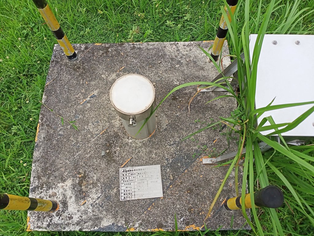A capped metal groundwater monitoring well head set in a concrete pad in grass, marked with a small information plate.