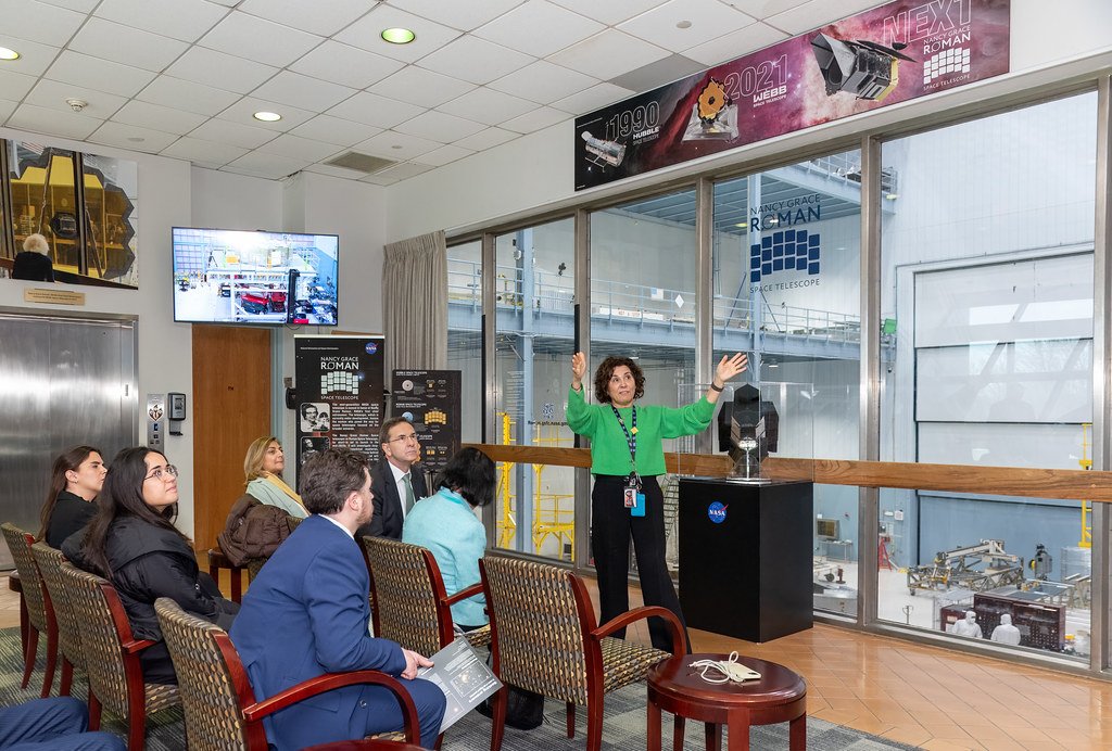 A NASA guide speaks to seated visitors in a gallery overlooking a clean room, with Roman Space Telescope signage on the windows.
