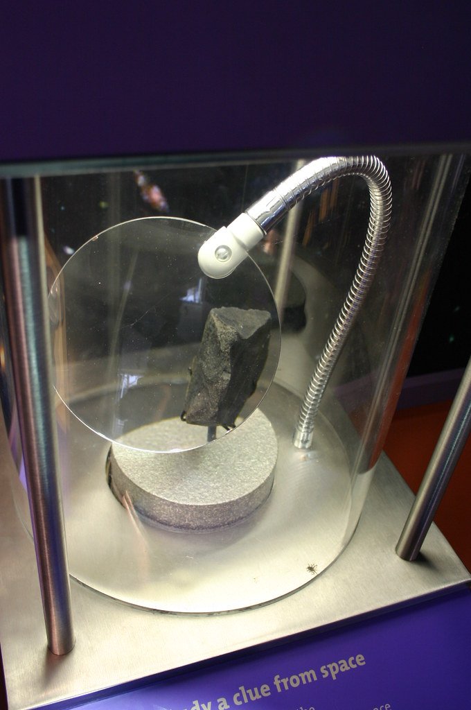 A small piece of the Murchison meteorite mounted under a magnifying lens inside a glass museum display case.