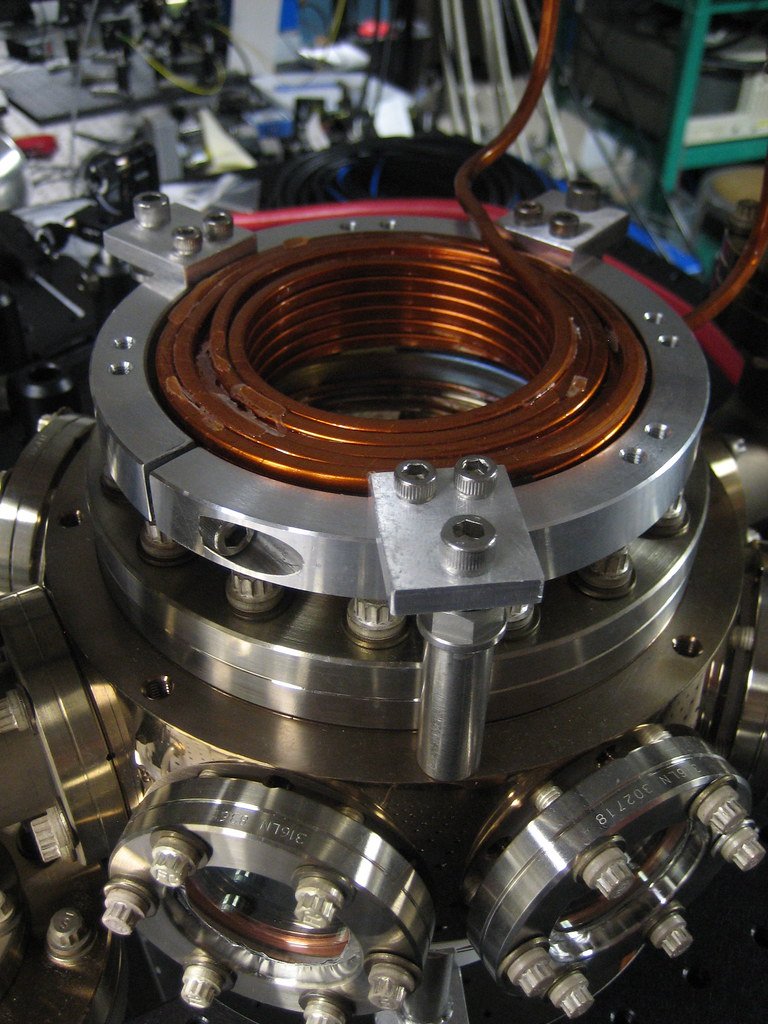 A close view of a copper magnetic coil mounted on top of a vacuum chamber ringed with bolted viewports.