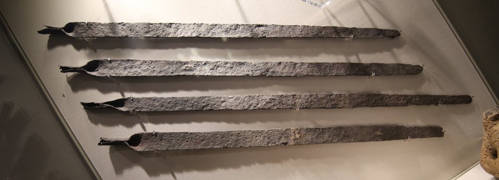 Four long, heavily corroded iron bars mounted side by side in a museum display case