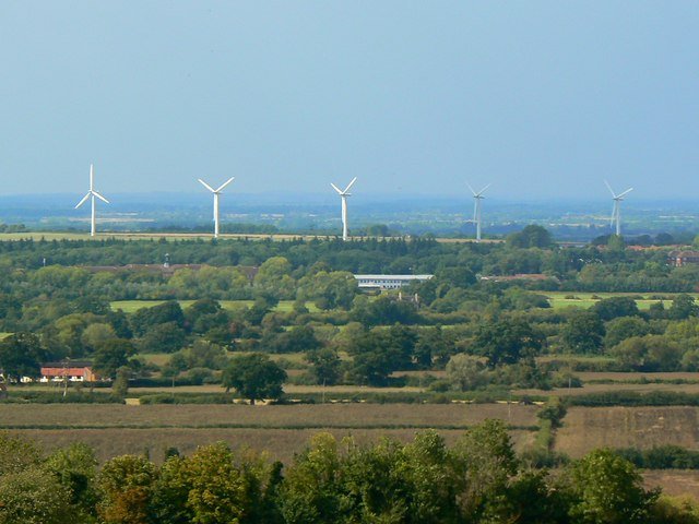 A wide view over English farmland with five wind turbines on the skyline and the long white line of a solar farm in the middle distance.