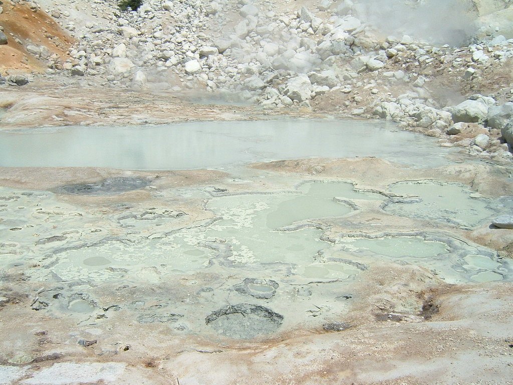 Steam drifting over shallow milky-green thermal pools and small mud pots in a pale mineral crust, with a rocky slope behind