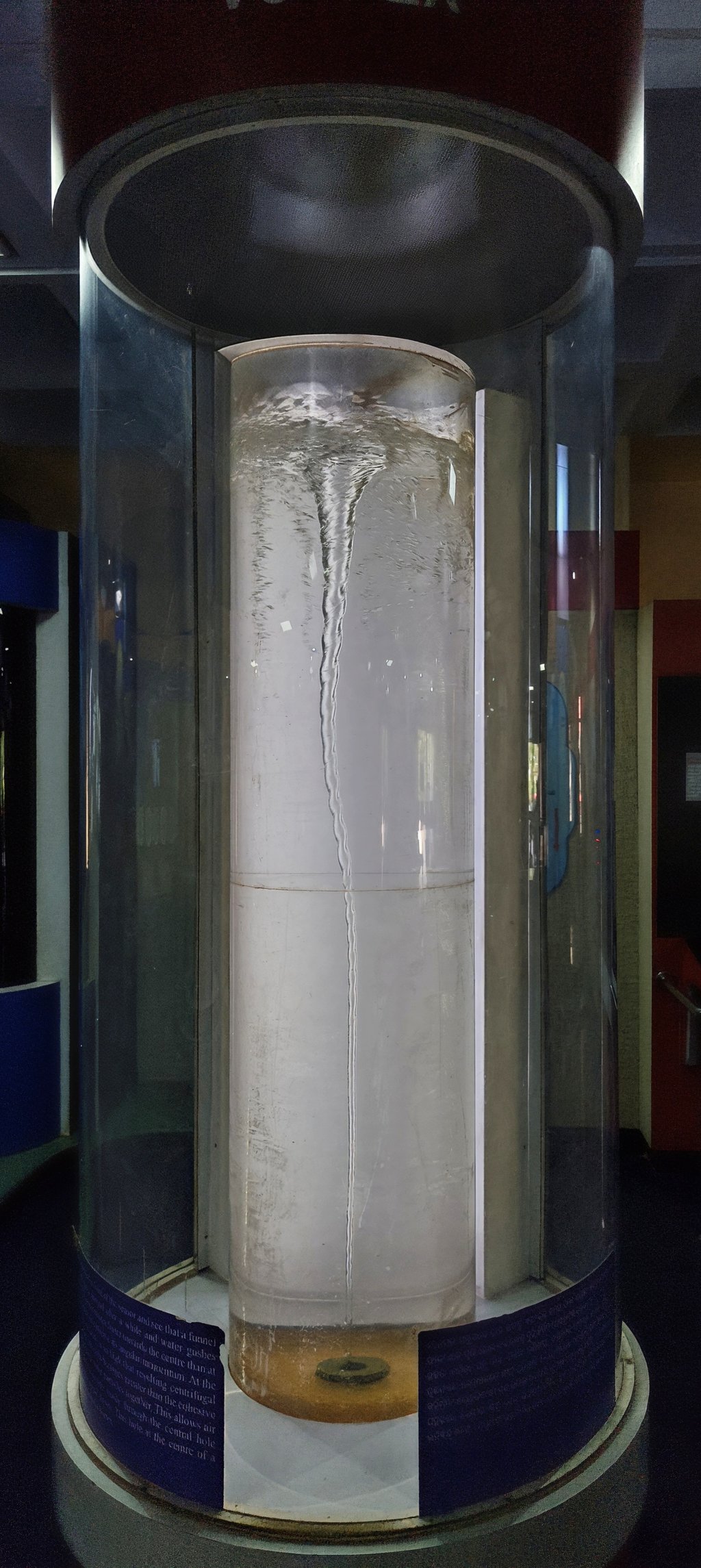 A tall glass cylinder of water in a science-center exhibit, with a thin twisting vortex running down its center.