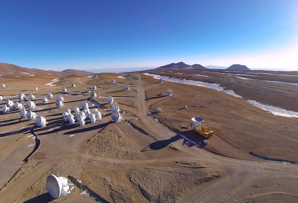 Dozens of white radio dishes spread across a high desert plateau under a deep blue sky, with volcanic peaks on the horizon.