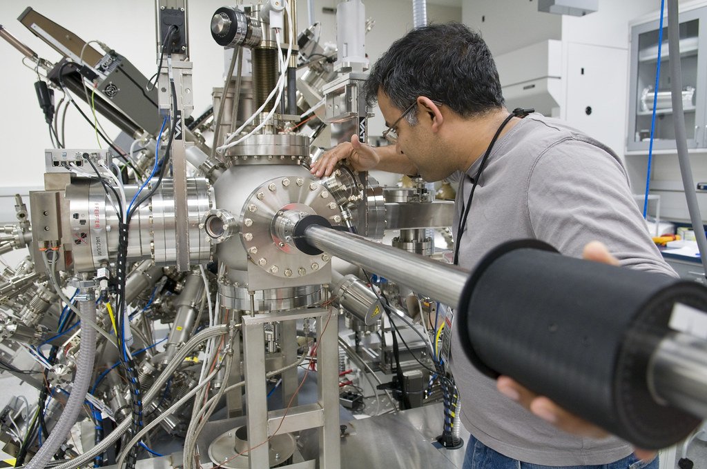 A researcher leans over a stainless steel molecular beam epitaxy chamber, reaching toward a port on the vessel.
