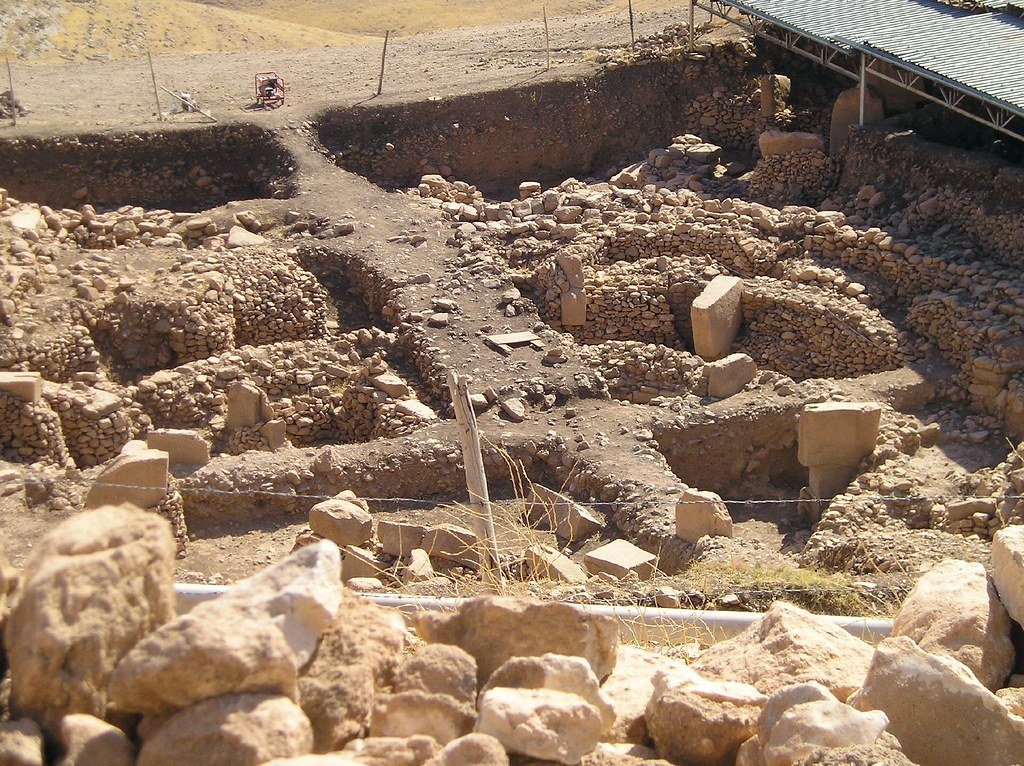 Open excavation trenches cut into the slope of the Gobeklitepe mound, with dry-stone walls, standing pillars and a shelter roof along the upper edge.