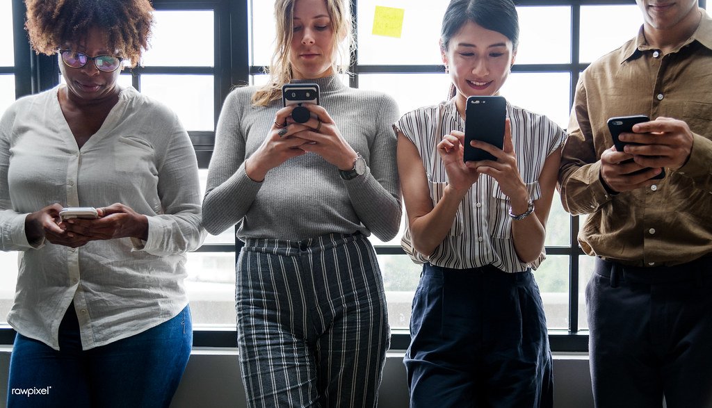 Four people standing side by side against a wall, each looking at a smartphone.