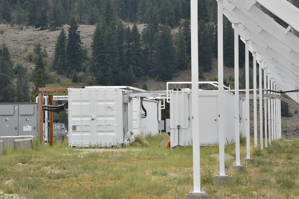White equipment containers lined up beside the telescope structure at the foot of a forested hillside.