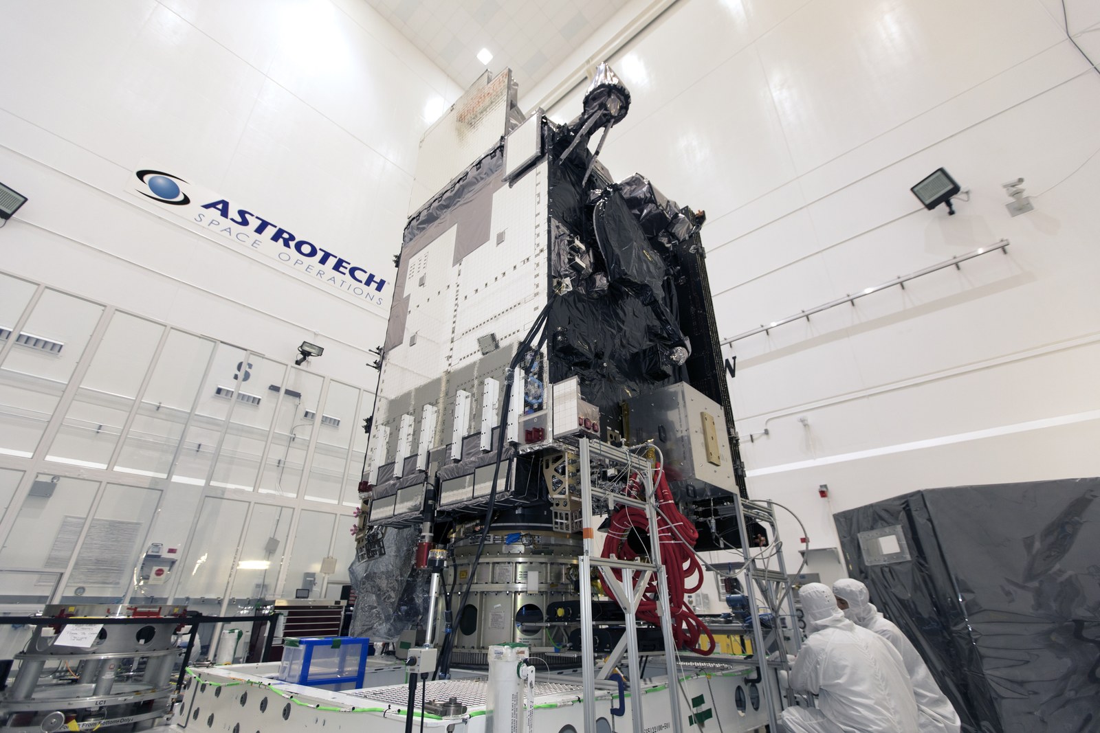 A tall boxy weather satellite stands upright inside a clean room while two technicians in white suits work beside it.