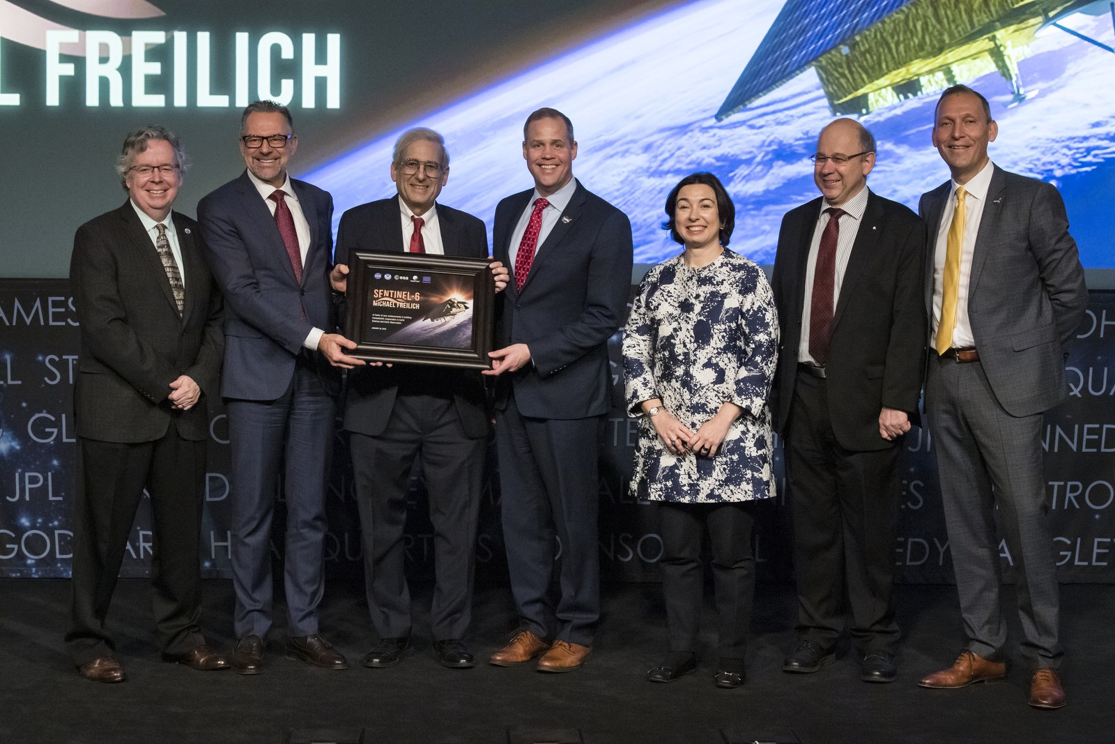 Seven people in formal dress stand on a stage, two of them holding a framed Sentinel-6 Michael Freilich mission plaque.