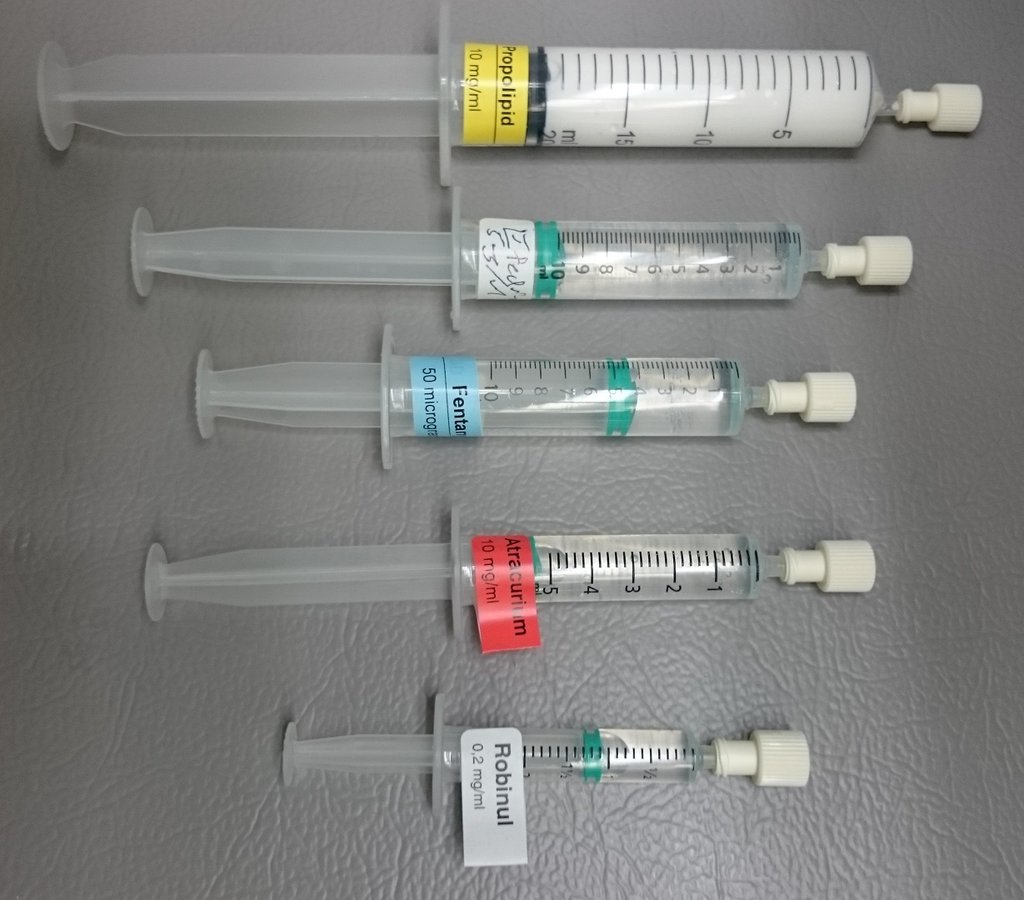 Five labeled syringes laid out in a row on a gray surface, drawn up with drugs used during anesthesia.