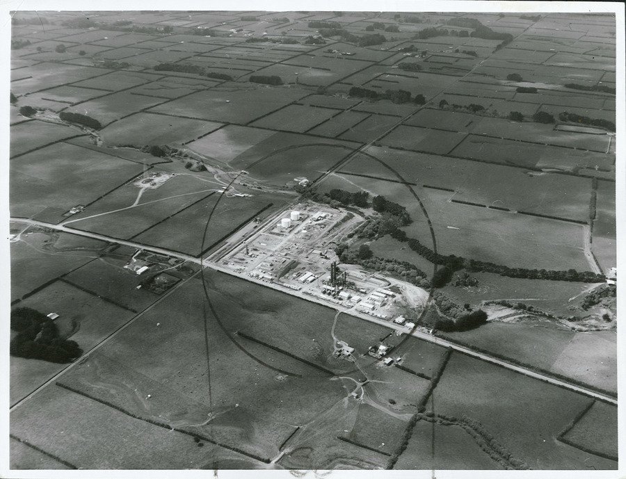 Black and white aerial photograph of an industrial gas site surrounded by a grid of farm paddocks, hedgerows and country roads.