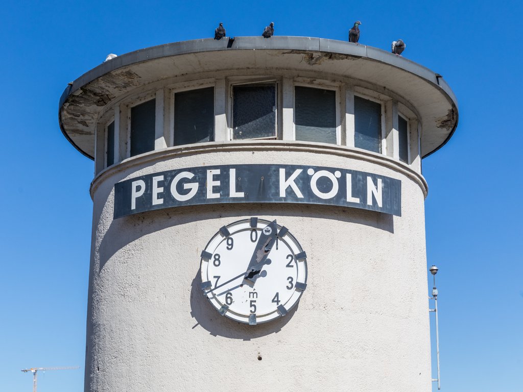 A white gauge tower on the Rhine bank in Cologne, Germany, lettered Pegel Koeln, with a large circular dial showing the river level.