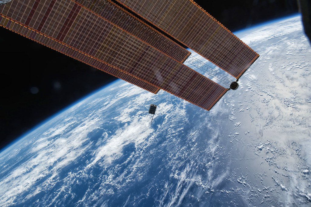 A small cube-shaped satellite drifts above the blue and white curve of Earth, with a space station solar array filling the upper left of the frame.