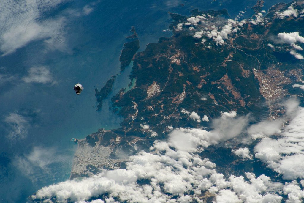 A small white and black capsule hangs above a cloud dappled coastline and blue ocean, photographed from the International Space Station.