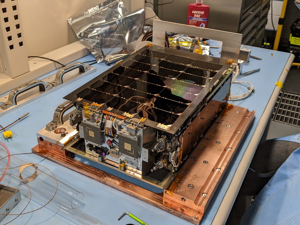 A compact spacecraft propulsion module with small thrusters, copper plumbing and a dark honeycomb panel sits on a laboratory bench.