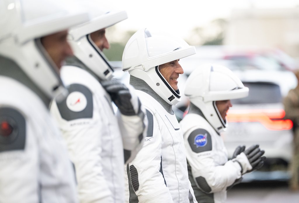 Four suited crew members walk in single file toward waiting cars, one of them smiling and another waving a gloved hand.
