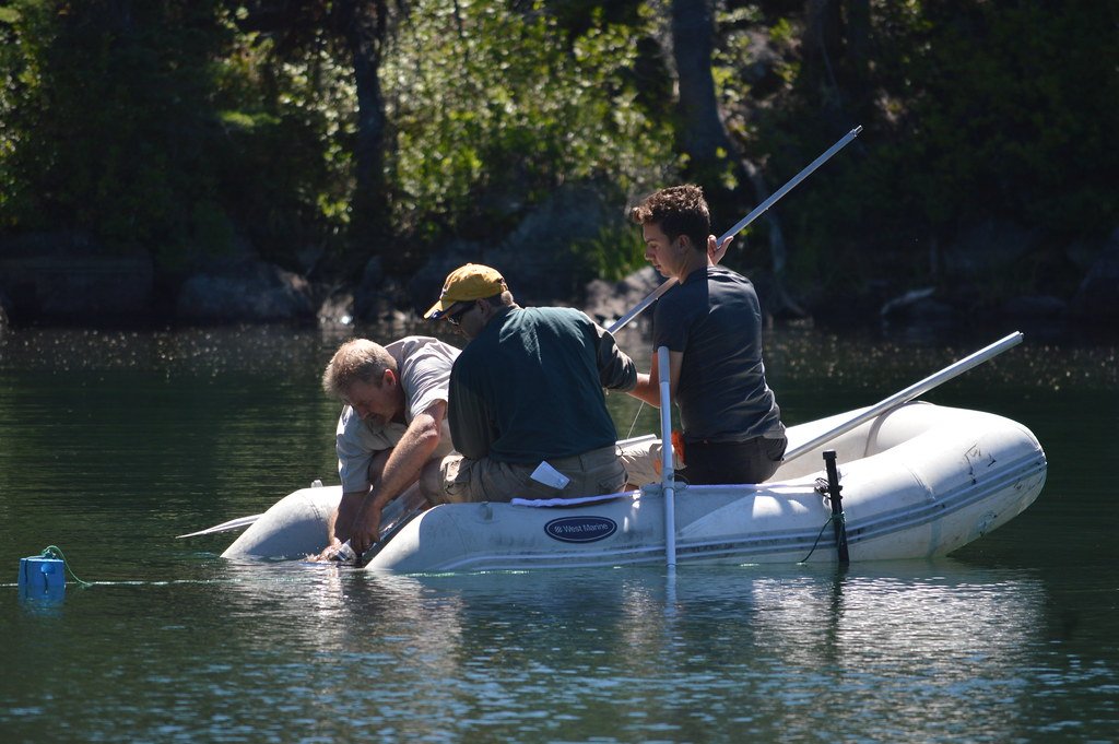 Three researchers in an inflatable boat lift a coring tube out of a calm forest lake.