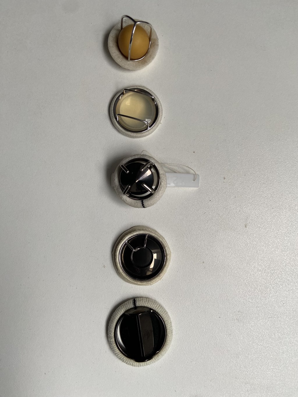 Five mechanical heart valve prostheses, each ringed by a fabric sewing cuff, laid out in a row on a gray surface.
