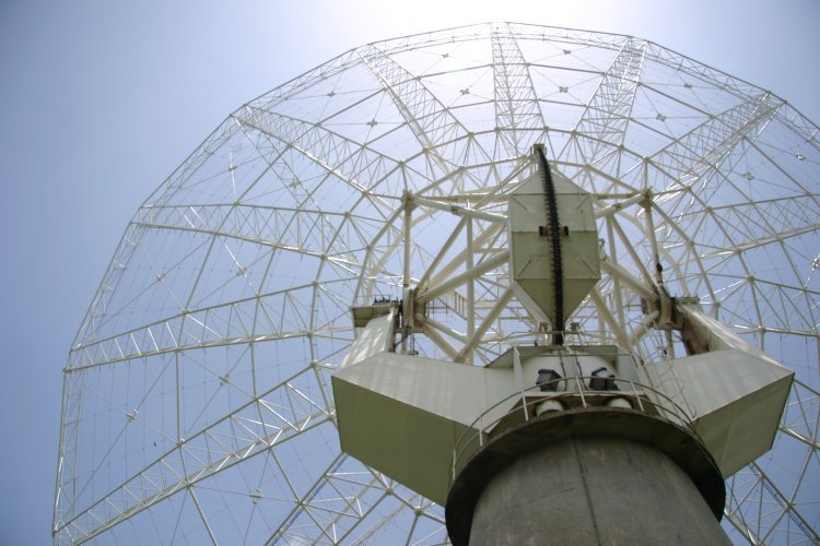 Looking up at the wire mesh dish of a large radio telescope against a bright sky.