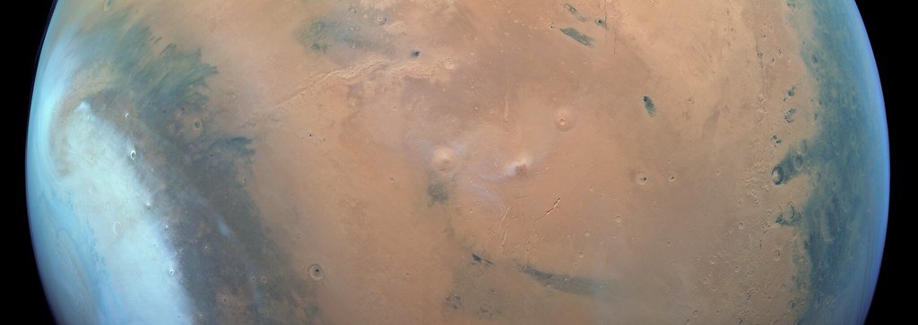 Wide orbital strip of ochre Martian plains with low volcanic domes near the center and bright hazes along the limb at left.