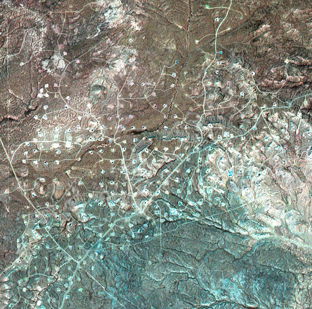 An aerial view in which pale well pads and the roads connecting them form a web across dry, hilly terrain.