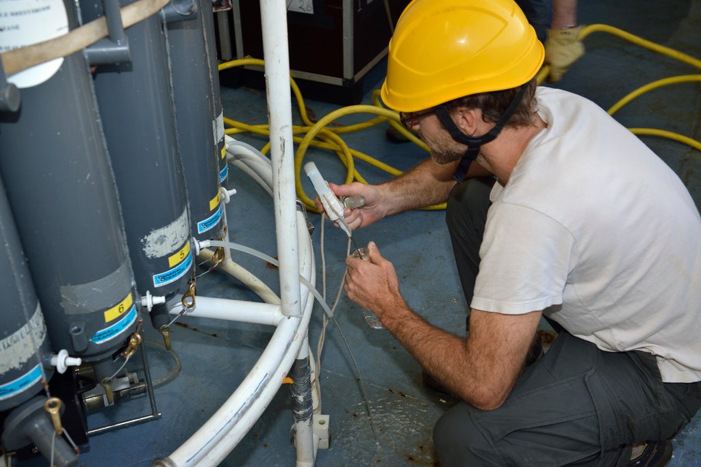 A crew member in a yellow hard hat crouches beside gray sampling bottles on a ship's deck, drawing water through a tube into a container.