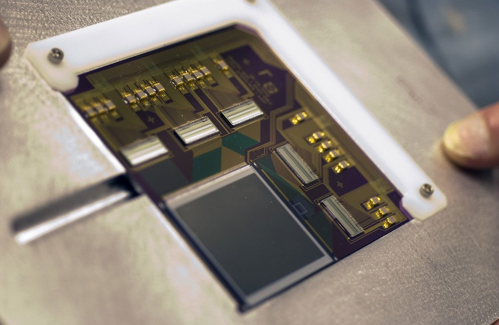 Close view of a small rectangular microshutter array chip held in a white frame, with gold and purple circuitry and a dark grid at its center.