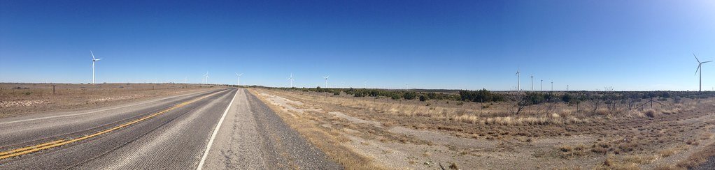 A wide view of a straight highway crossing flat grassland, with rows of turbines on the horizon under a clear blue sky.