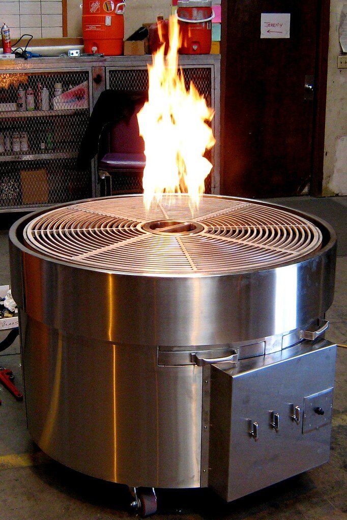 A large round stainless steel commercial gas broiler with a circular grate, flames rising from its center, standing on a workshop floor.