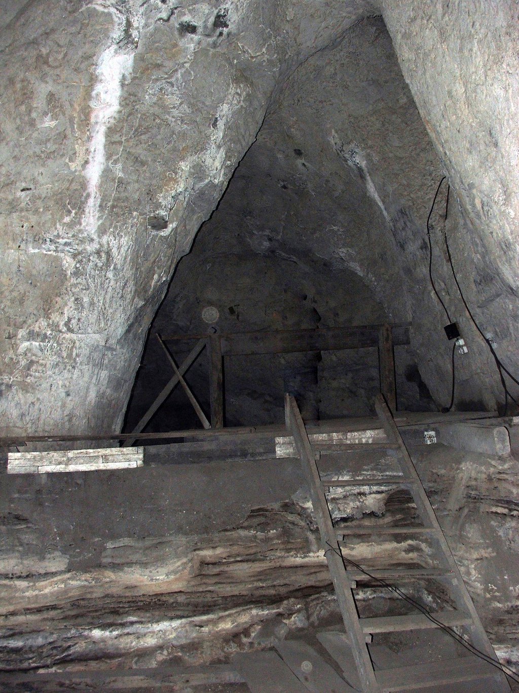 The inside of a cave chamber with bare rock walls, a wooden ladder and timber framing set against the excavated floor.