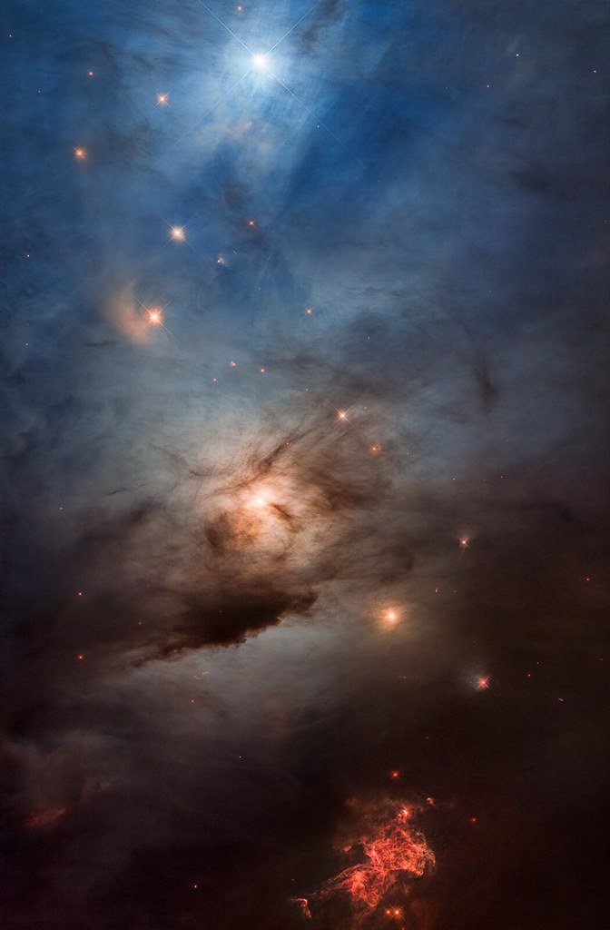 Wide view of a blue reflection nebula and dark dust lanes, with red knots of shocked gas at the bottom.