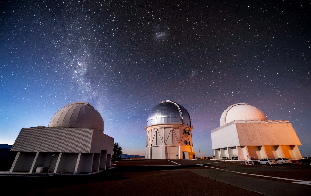 Three telescope domes on a Chilean mountaintop at night, with the Milky Way and the Magellanic Clouds above them.