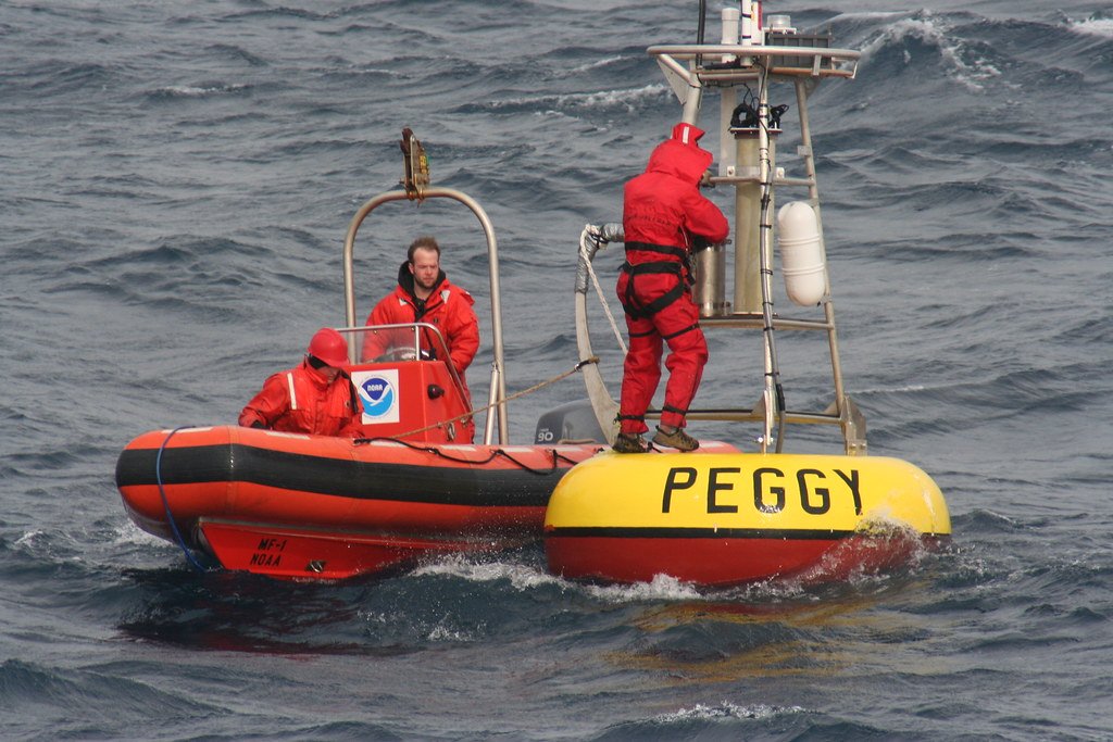 Two people in survival suits in an inflatable boat hold alongside a yellow and red moored buoy in choppy gray water while a third climbs its tower.