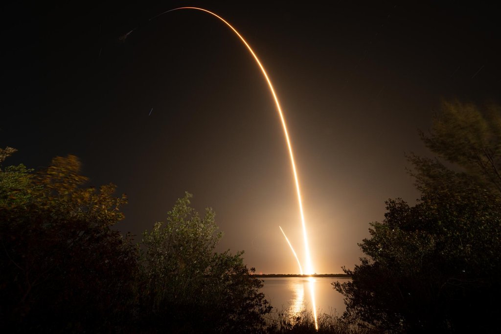 A long exposure shows a bright curving arc of rocket exhaust rising from the horizon into a night sky, mirrored in the water below and framed by dark trees.