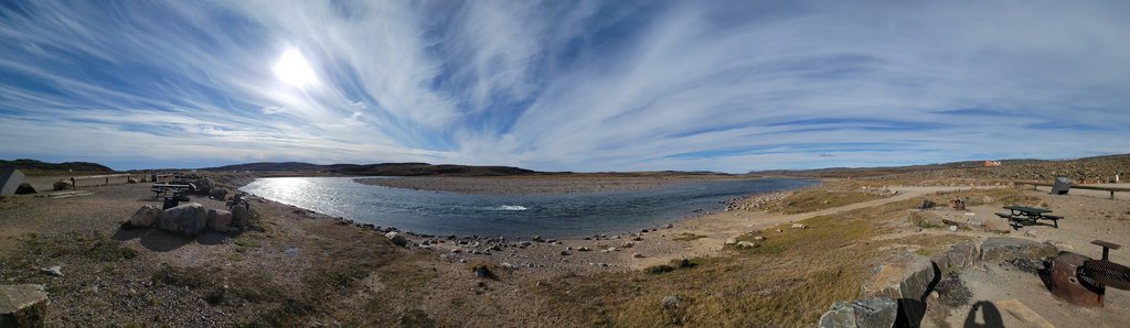 A wide panoramic view of a river mouth meeting the sea, with bare rock, tundra and a picnic area in the foreground.