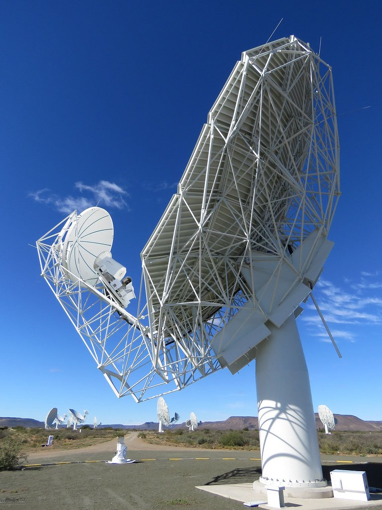A white radio dish of the MeerKAT array tilted toward a clear blue sky, with more dishes along a road in the distance.