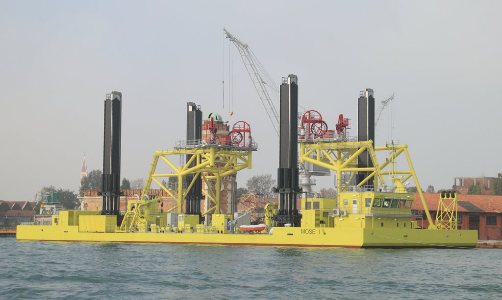 A yellow jack-up barge with four tall legs and deck cranes, lettered Mose I on the hull, moored on the water in Venice.