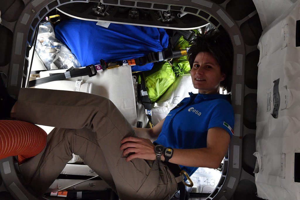 An astronaut floats inside a cargo freighter whose walls are lined with blue and green bags of packed waste and used equipment.