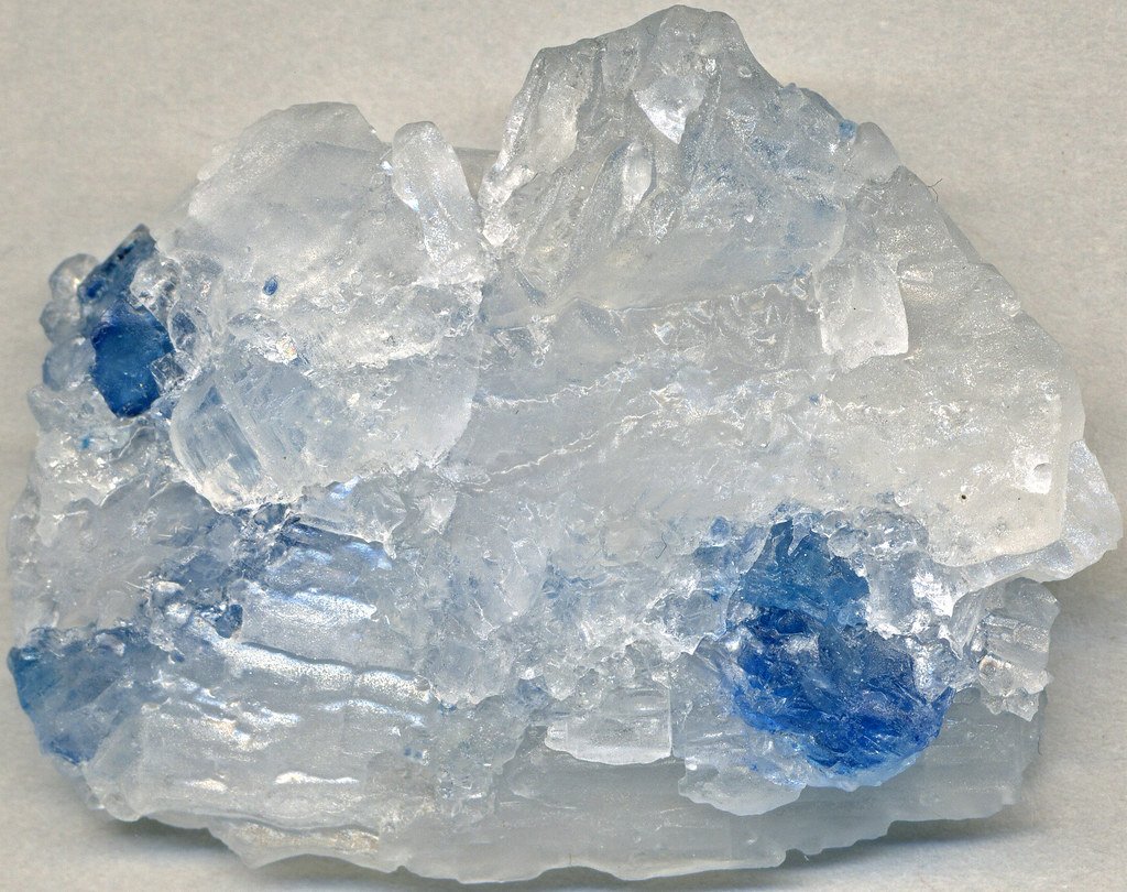 A chunk of translucent white rock salt with several patches of deep blue coloration, photographed against a pale background.